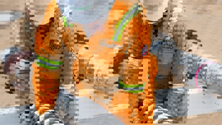 Engineer in orange Welltec coveralls and white helmet working on large metallic pipes at an outdoor industrial site.