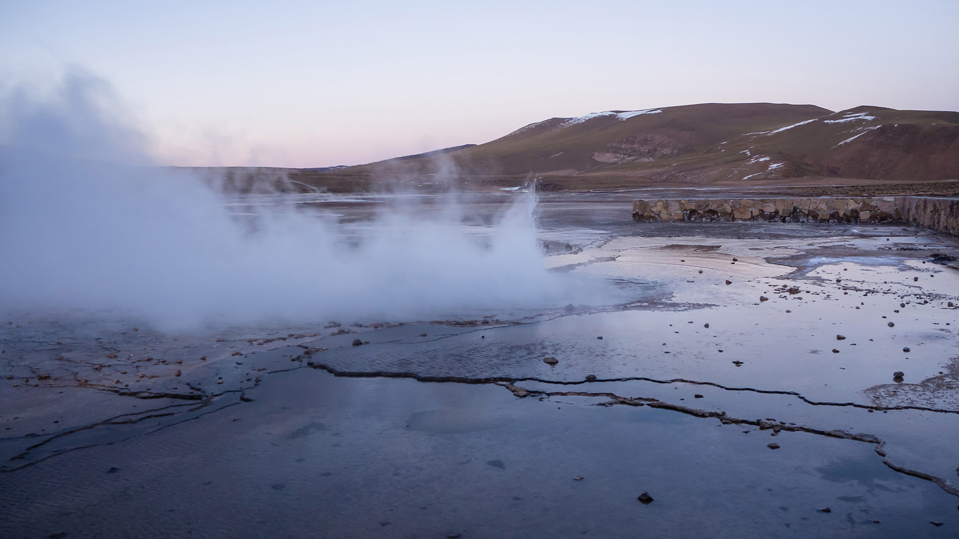 Geothermal field with steam rising from the ground, surrounded by rocky terrain and snow-capped hills in the distance.