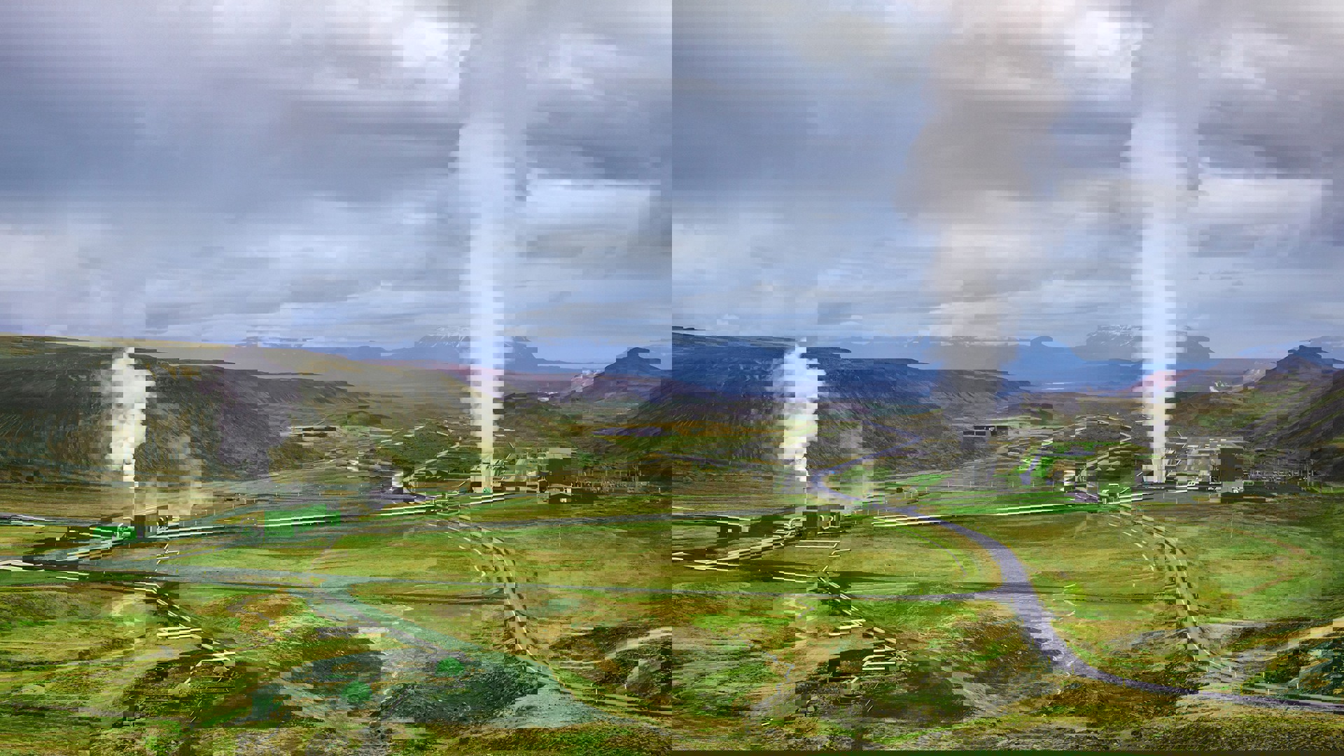 A large green field with visible steam emerging from the ground, suggesting geothermal activity in the area