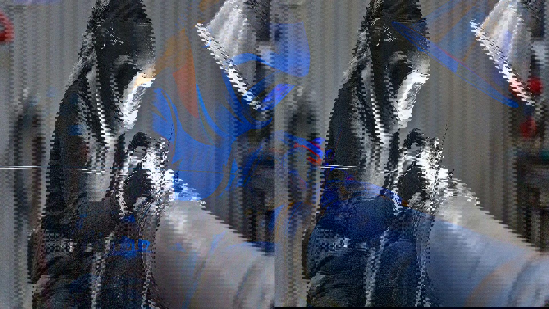 Woman welding a metal pipe with a protective helmet and gloves, illuminated by bright light in a workshop setting.