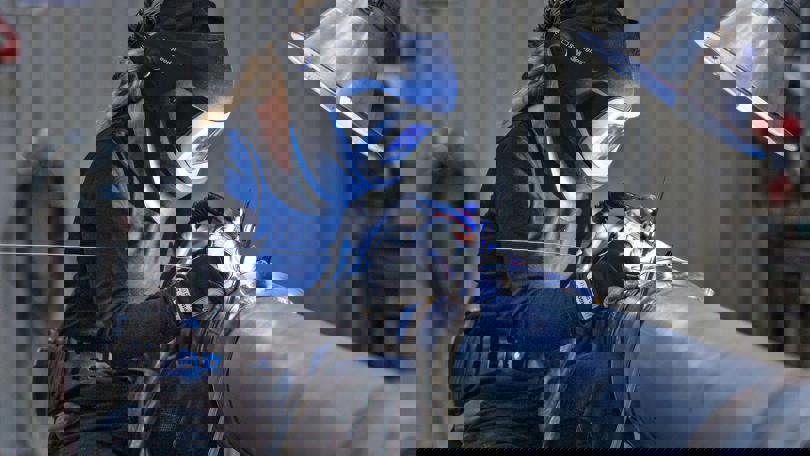 Woman welding a metal pipe with a protective helmet and gloves, illuminated by bright light in a workshop setting.