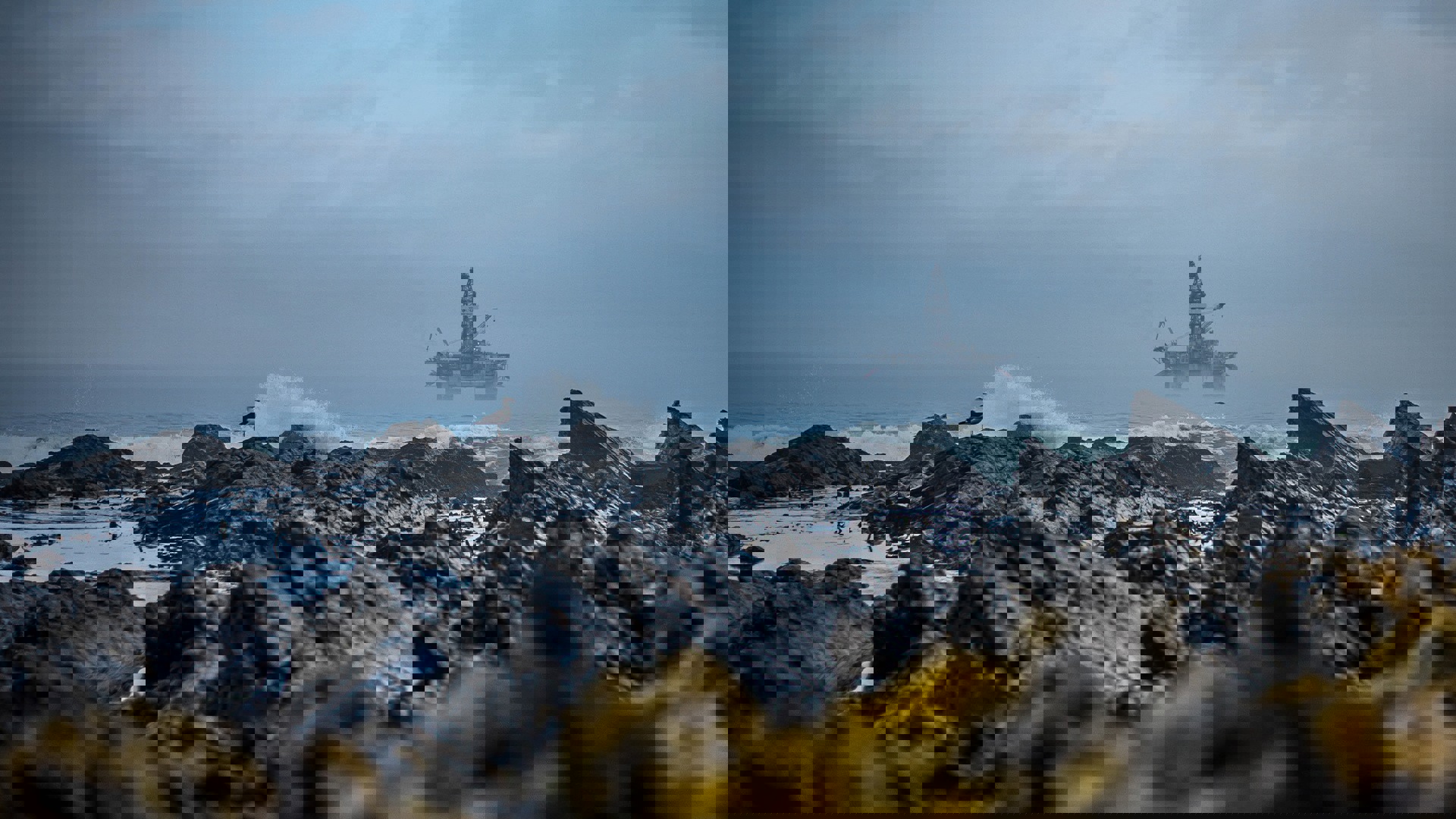  Offshore oil rig in the distance with rocky coastline in the foreground, waves crashing, and a seagull perched on a rock under a cloudy sky.
