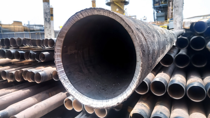 Close-up view of a large, cut-open production pipe with stacks of similar pipes in the background at an industrial workover site.