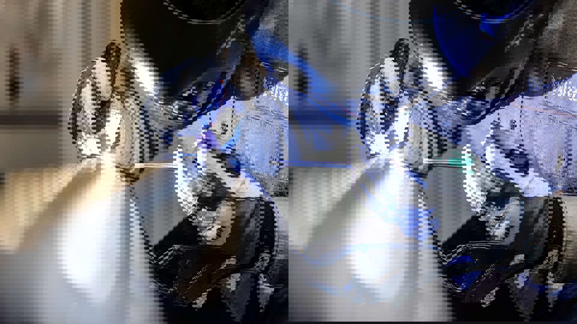 A man wearing a welding helmet is focused on welding a metal pipe in a workshop setting.