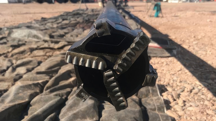 Downhole tool on rig floor, positioned upright in front of a drilling rig structure, captured in sunlight with surrounding equipment visible.