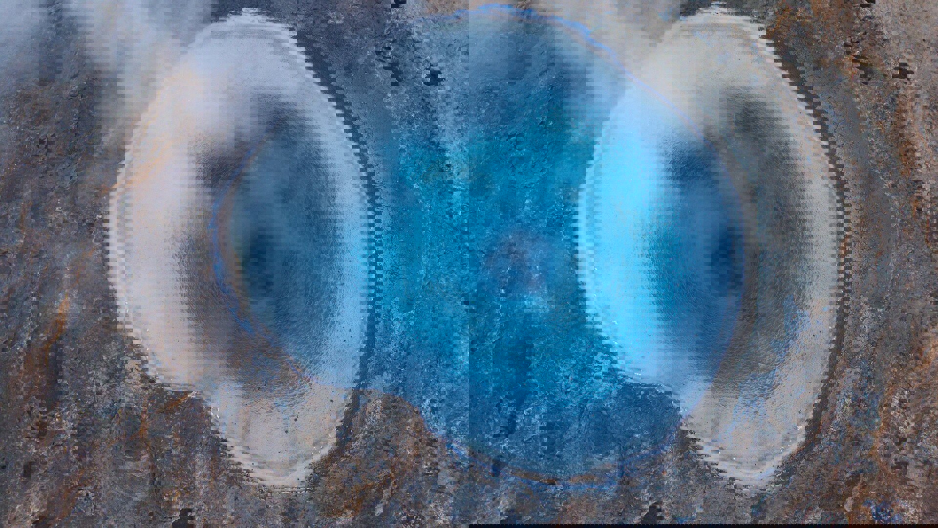 A blue geothermal hole in the ground releases steam, highlighting its unique color and natural surroundings.